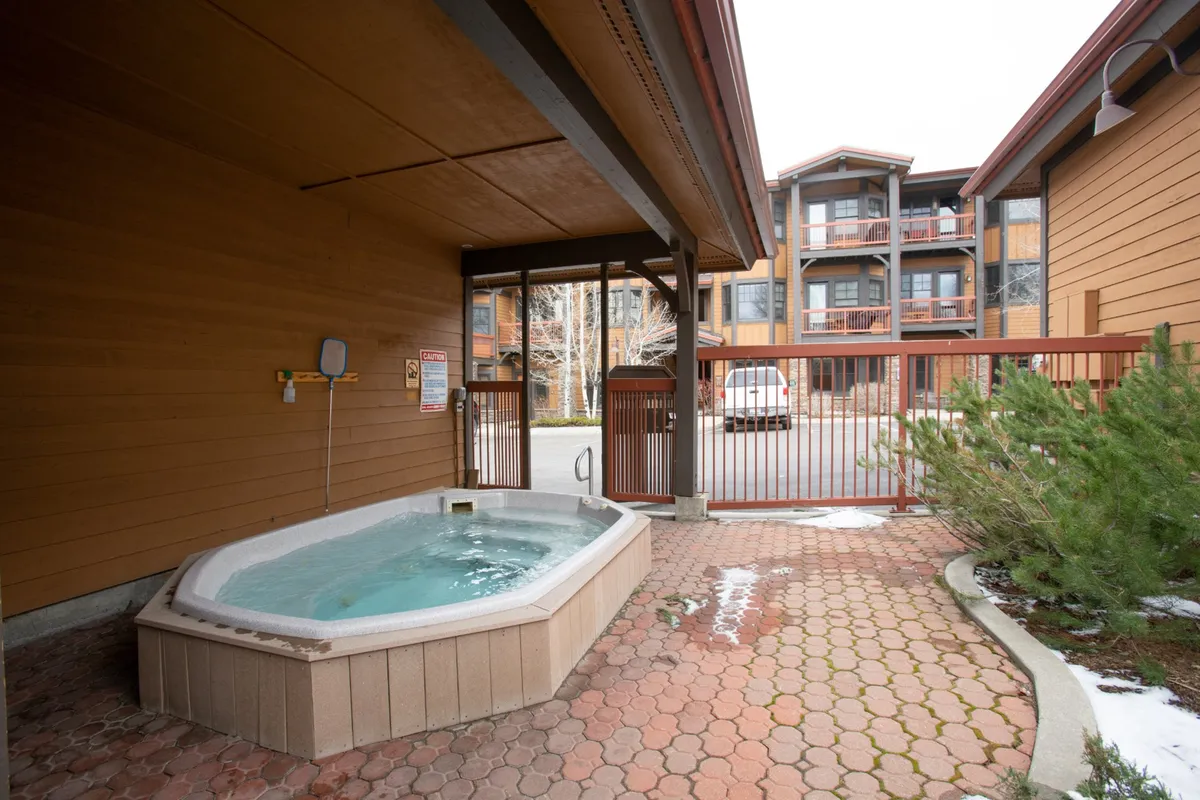 An exterior view showcasing the hot tub area with surrounding architecture and a paved pathway. The atmosphere is inviting and social, with ample space for guests to gather and enjoy the outdoors. Evening lighting hints at the possibility of starlit soaks.