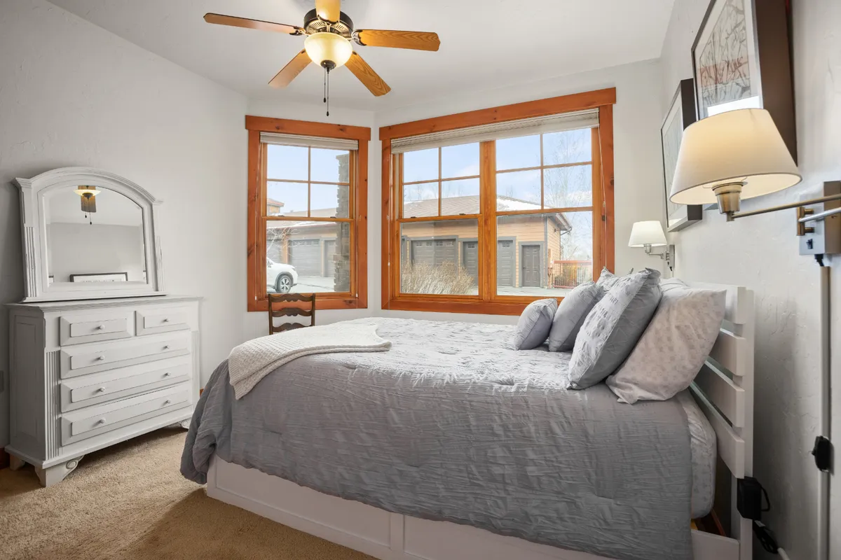 A serene bedroom with gray bedding, large windows looking out to the landscape, a white dresser, and a ceiling fan. The natural light streaming in suggests an ever-changing view and emphasizes the home's restful qualities after a day of seasonal exploration.