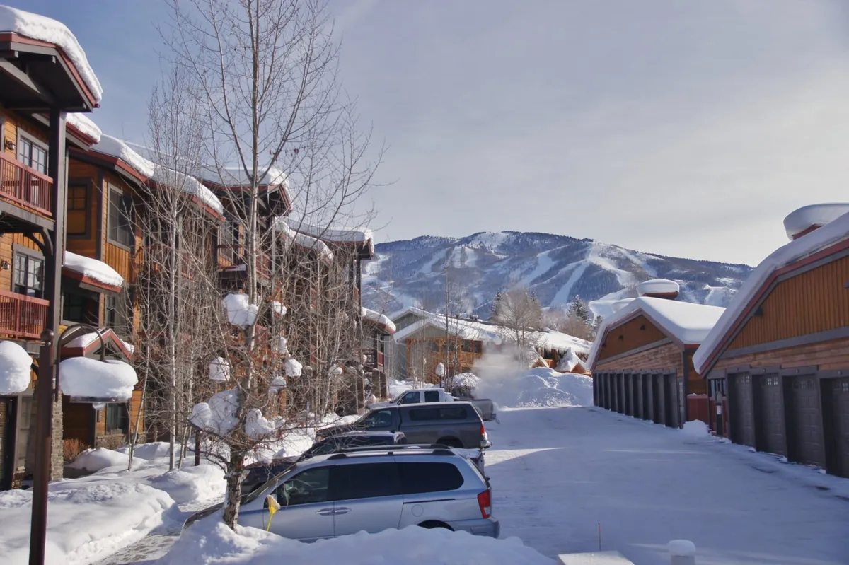 A tranquil winter scene features snow-covered rooftops and tree branches, vehicles in the parking lot, and a backdrop of mountains. The image evokes a feeling of arrival at a cozy mountain retreat after a day of outdoor exploration.