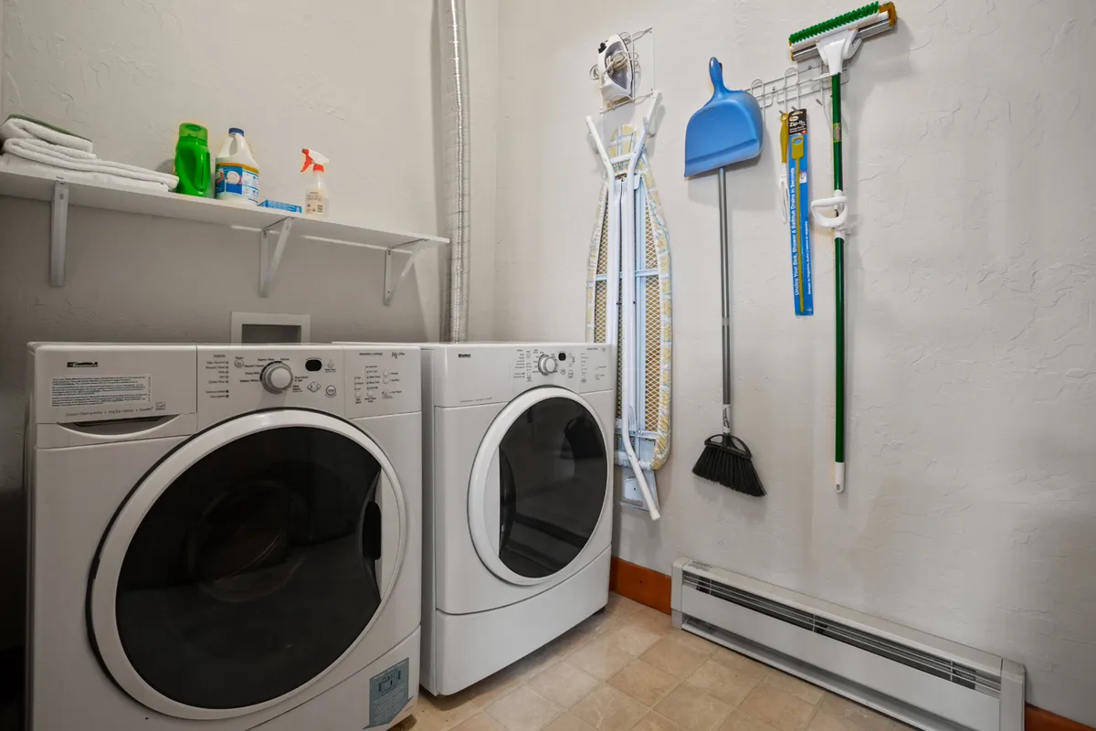This image features a dedicated laundry area equipped with a washing machine, dryer, cleaning supplies, and an ironing board. The space is tidy and organized, clearly set up for practical, in-unit laundry tasks.
