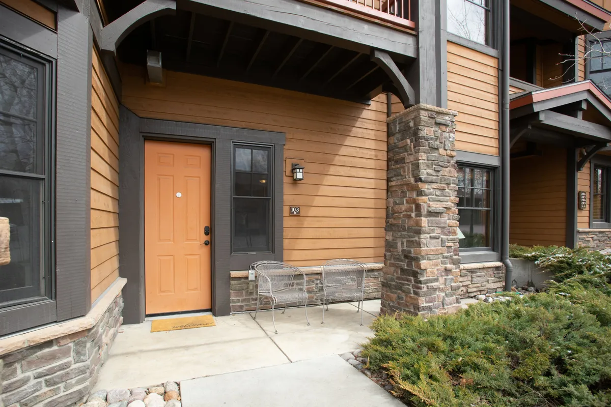 The image shows a welcoming porch with an orange door, stone pillar, metal chairs, and lush green shrubs. The setup suggests a comfortable spot for families to sit outside, enjoy fresh air, and connect with the neighborhood. The entryway feels accessible and encourages outdoor relaxation.