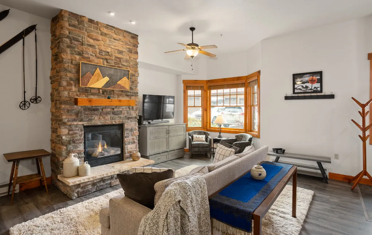 The image shows a cozy and stylish living area with a beautiful stone fireplace as the centerpiece, inviting bay window seating perfect for relaxing, contemporary sofa and coffee table, and tasteful decorative wall art. The scene is warmly lit, creating a welcoming and comfortable atmosphere ideal for unwinding after a day in the mountains.