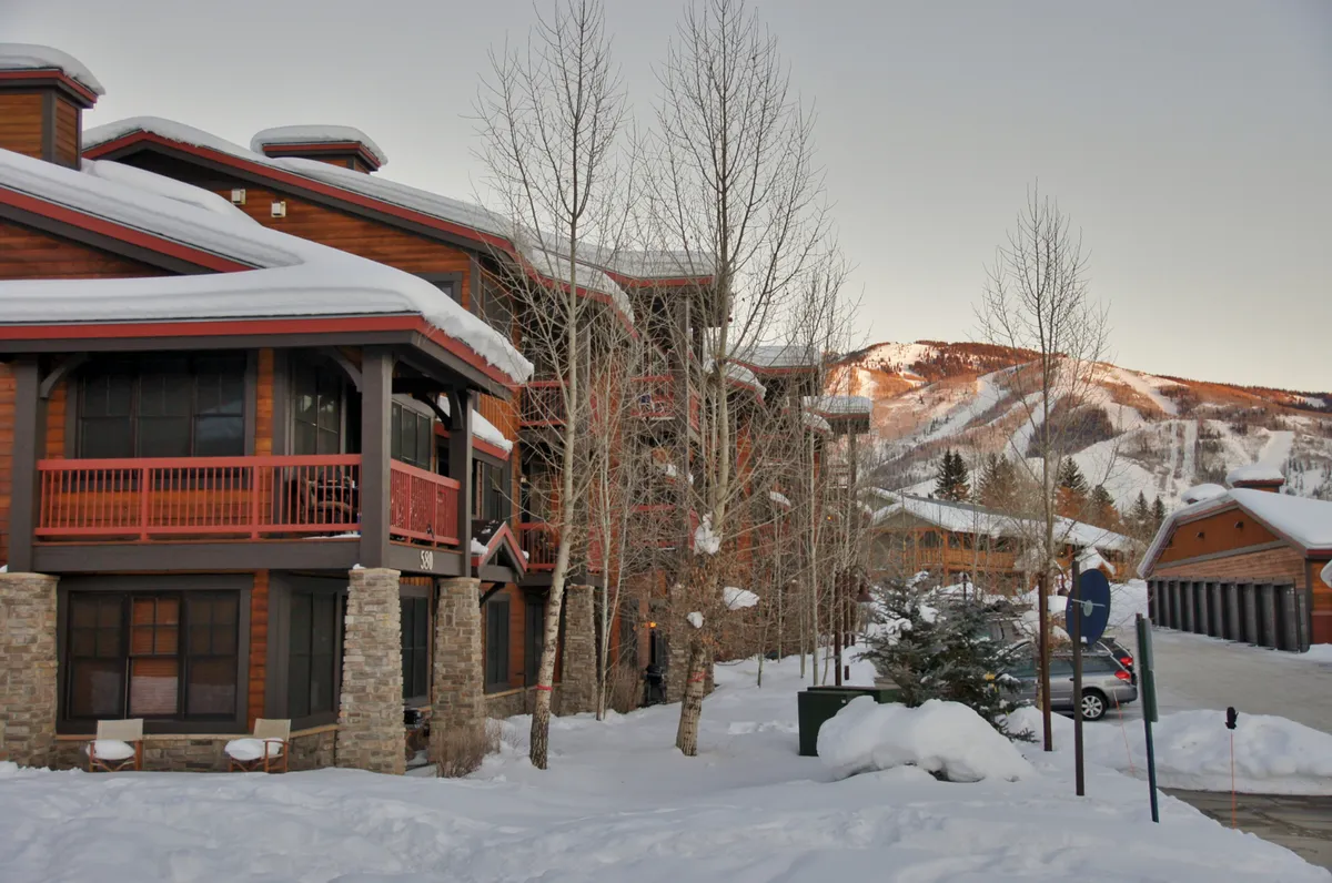 A view of wooden architecture surrounded by a snow-blanketed landscape, with distant mountains and aspen trees. Balconies suggest spaces for relaxing outdoors, watching sunsets, or enjoying the mountain air after a day of adventure.