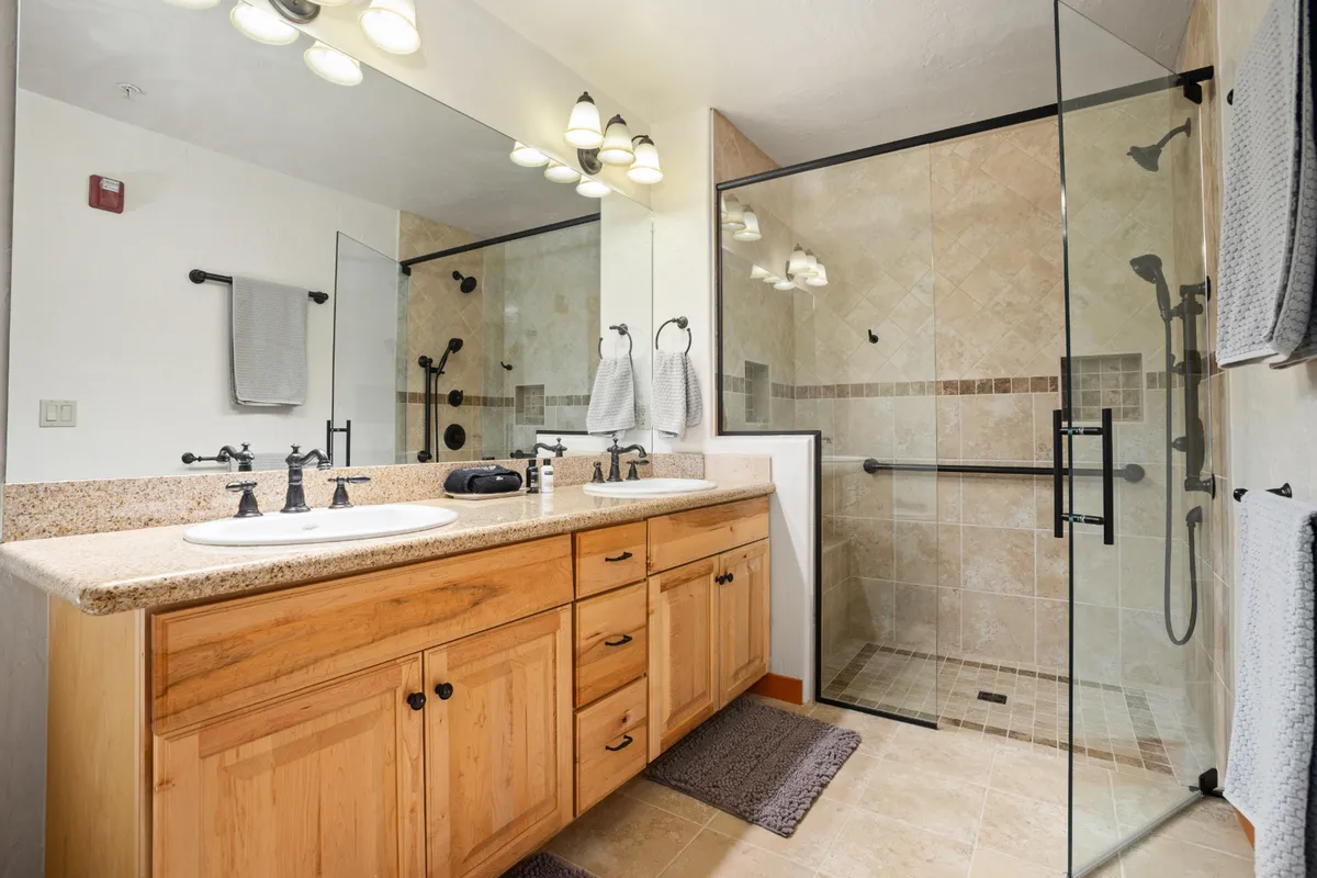 A modern bathroom with a double vanity sink, a glass-enclosed walk-in shower with a bench, granite countertops, and wood cabinetry. The space is bright and inviting, designed for comfort and self-care.