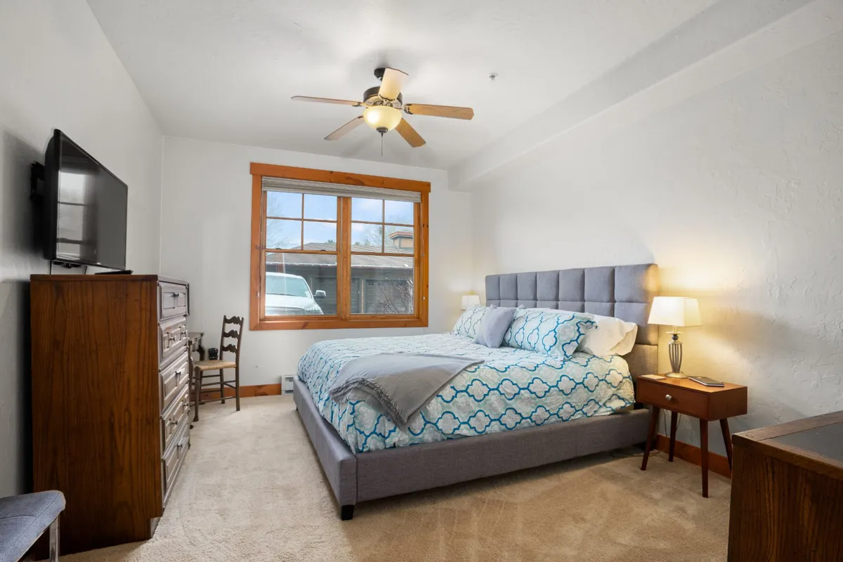 A sunlit bedroom featuring a comfortable bed with patterned bedding, a wooden dresser, a large window draped with blackout curtains, and a ceiling fan overhead. The space exudes tranquility, with plush bedding and natural light filtering in to create a restful atmosphere.