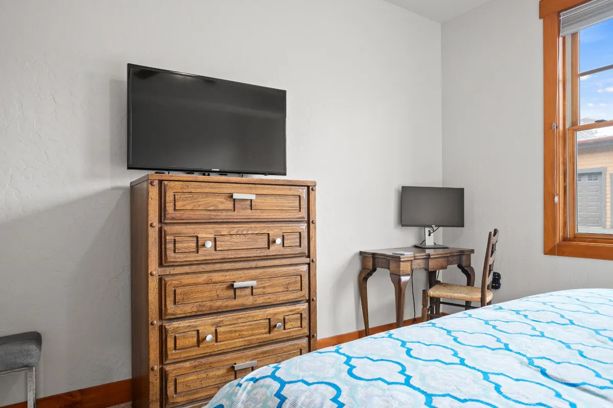 A cozy bedroom with a wooden dresser, a small desk equipped with a monitor, and a bed adorned with a patterned cover. The window provides ample natural light, and the setup is ideal for both relaxation and productivity.