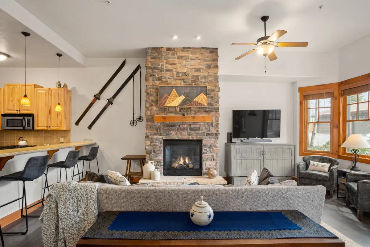 This image captures a welcoming open-plan living space featuring a stone fireplace, a comfortable sofa with a decorative throw, a kitchen bar, and mountain artwork accenting the walls. The scene is warm and lively, suggesting a perfect gathering spot after a day out.
