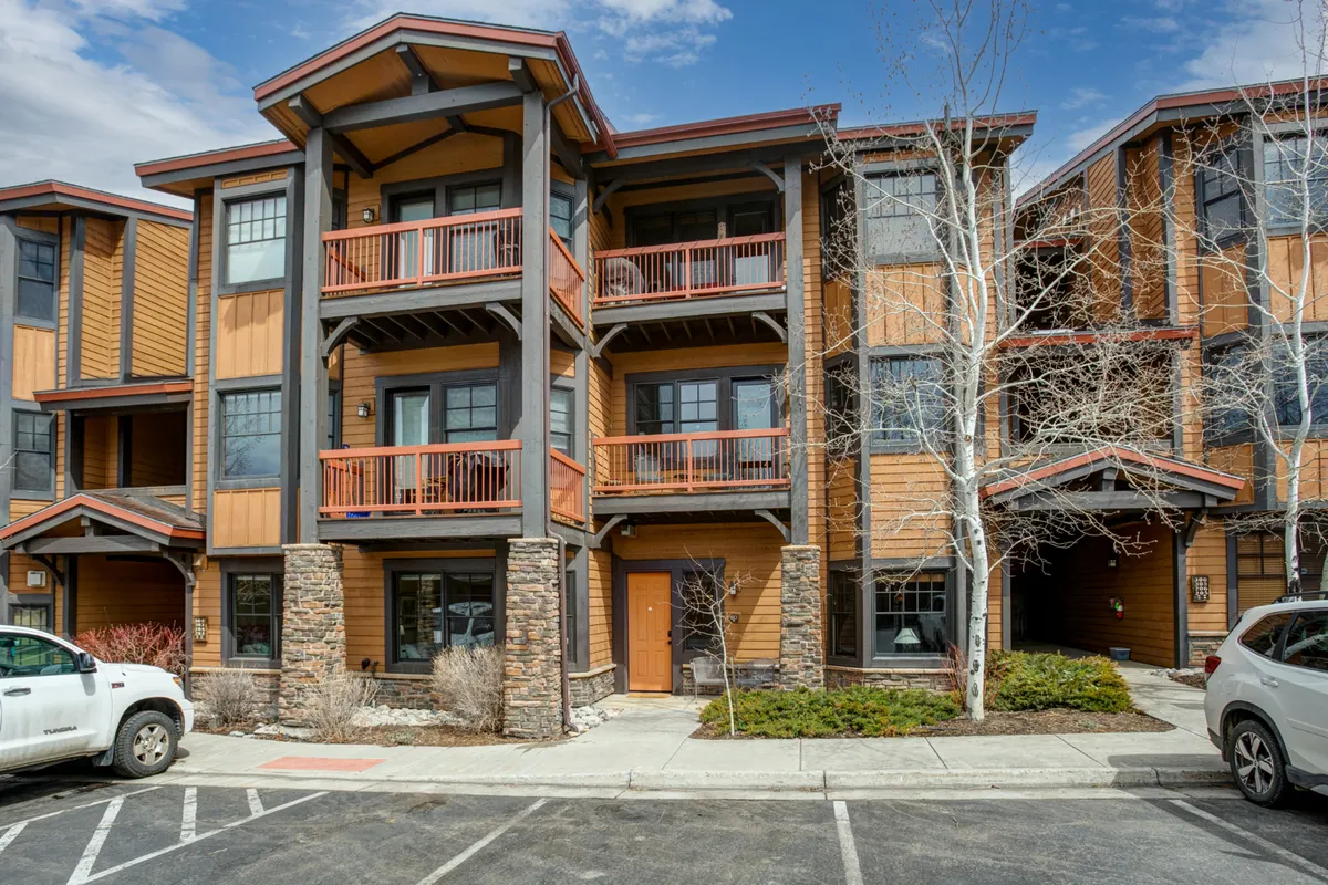 A wide exterior shot featuring wooden balconies, stone accents, large windows, and a tree-lined pathway. The scene is cozy and inviting, with hints of people enjoying the balconies and walking amidst nature, evoking a vibrant community vibe.