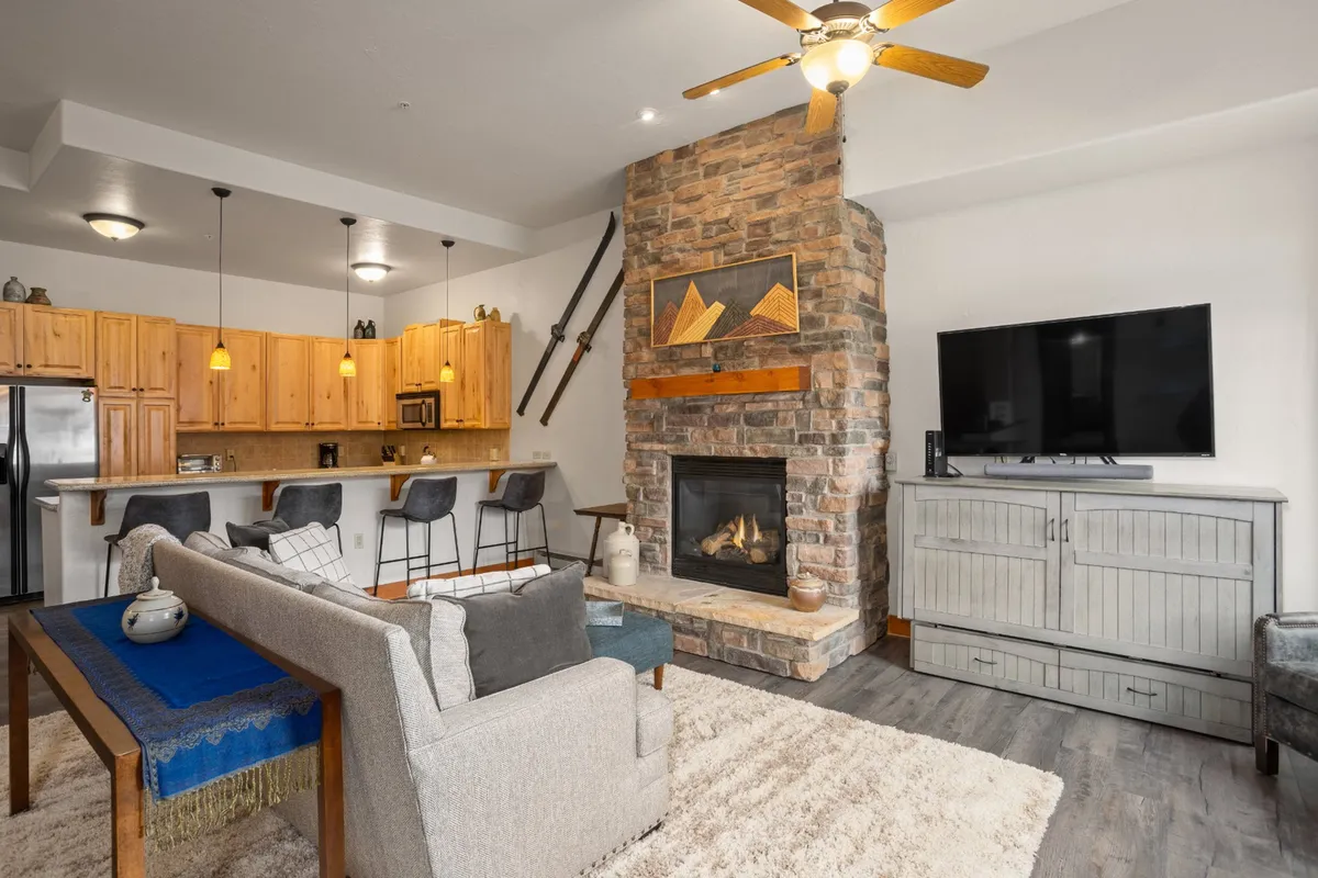 This inviting scene features a large stone fireplace, a comfortable seating area, visible kitchen cabinetry, and a television. The open-plan design highlights spaces for cooking, relaxing, and entertaining guests, linking together multiple communal activities.