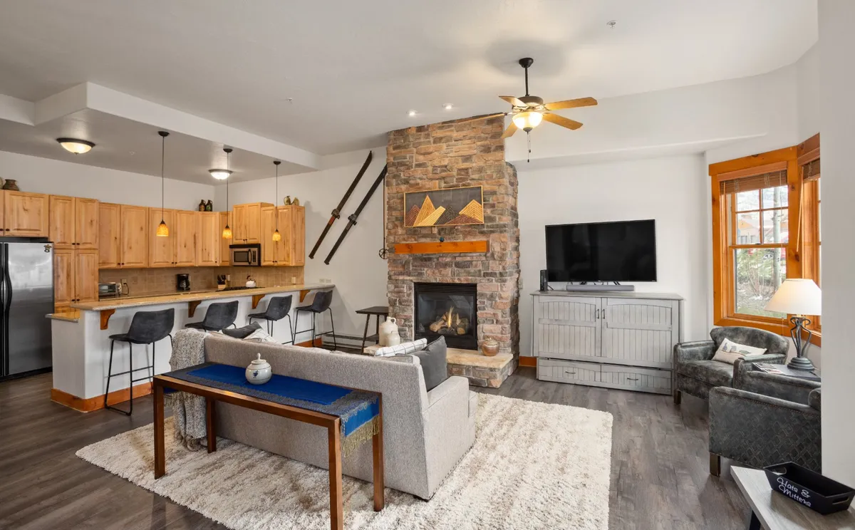 A wide-angle view of an open-concept living area featuring a stone-framed gas fireplace, a chic sofa, leather chairs, a large SmartTV, and a kitchen island in the background. Natural light streams in through generous windows, illuminating both the living and kitchen areas. The Murphy bed cleverly blends into the TV stand, showcasing the room's flexible layout.
